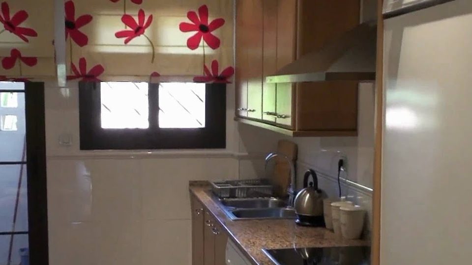 Frame from the apartment tour video: the kitchen, with a red flower-print blind, sink, kettle and wooden cupboards